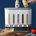 The Cereal Dispenser with its 5 grids full, resting on a wooden surface In front of a blue wall, surrounded by condiments. A human hand resting sauce jars on top of the dispenser.