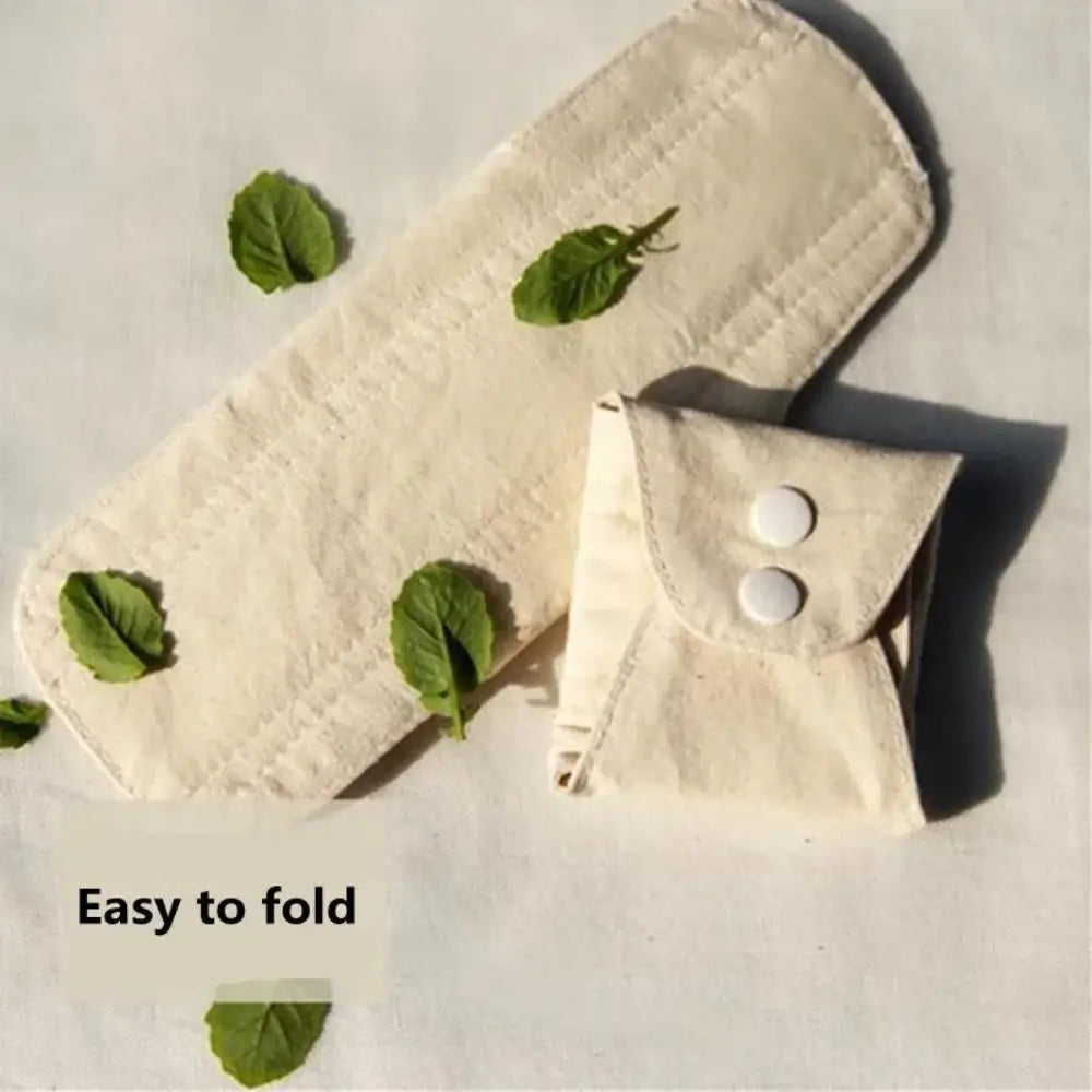 An opened Reusable Sanitary Pad and a folded, buttoned up pad next to each other with tiny leaves resting on and around the pad.